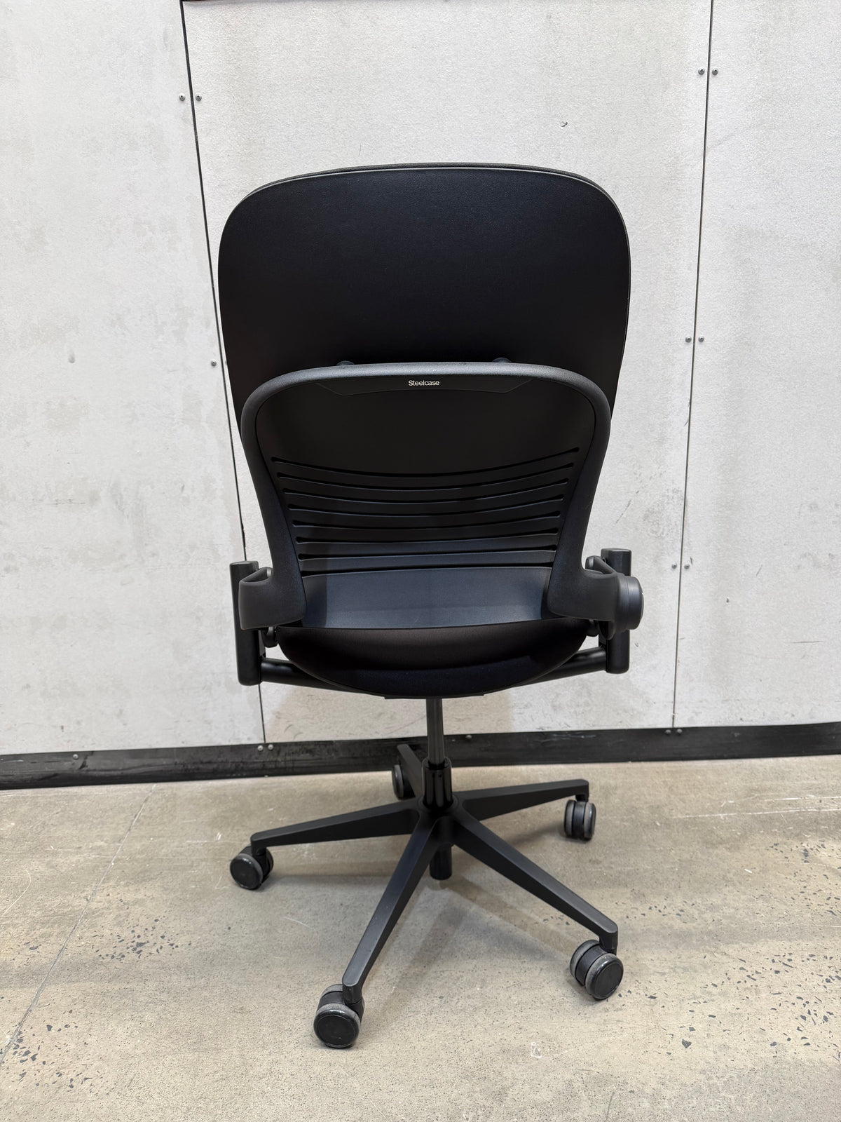 A black ergonomic office chair with a high curved backrest, mesh back support, adjustable armrests, and a five-wheel base, photographed against a light gray paneled wall and concrete floor.