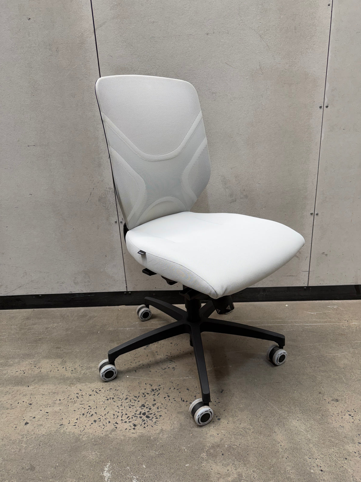 A modern white office chair with a curved ergonomic backrest, white padded seat, and a black five-star wheeled base, photographed against a gray concrete wall and floor.