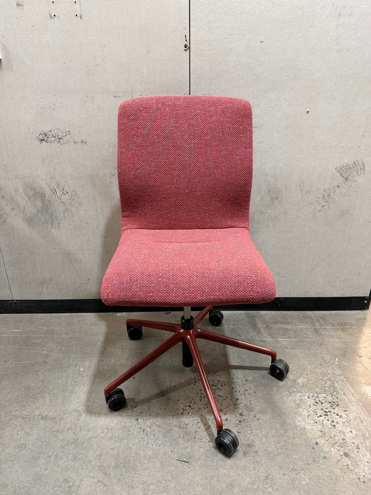 A red upholstered office chair with a five-star metal base and black caster wheels, photographed in front of a concrete wall and floor.
