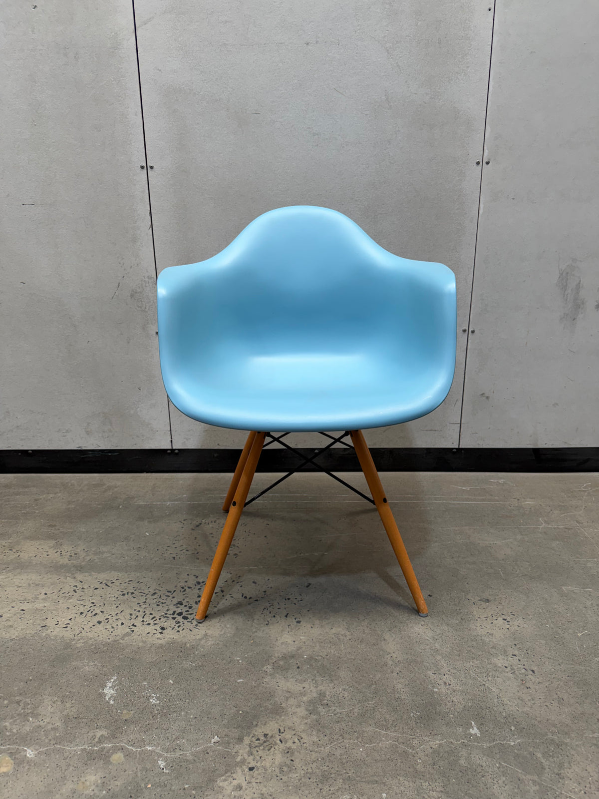 A light blue molded plastic armchair with tapered wooden legs, photographed in a warehouse or showroom setting against a gray concrete wall and floor.